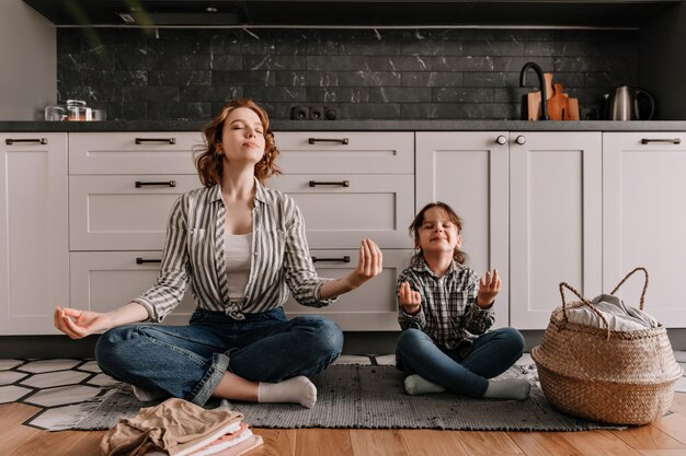 Mamá pacificada y su pequeña hija están sentadas en el piso de la cocina y meditan con una sonrisa