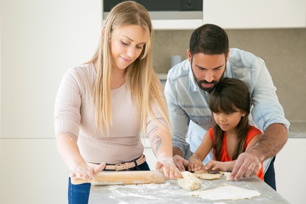 Mamá, papá e hija amasando y enrollando masa en la mesa de la cocina con harina en polvo.