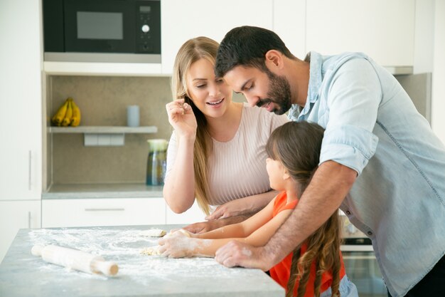 Mamá y papá enseñando al niño a amasar la masa en la mesa de la cocina con harina desordenada. Pareja joven y su chica horneando bollos o pasteles juntos. Concepto de cocina familiar