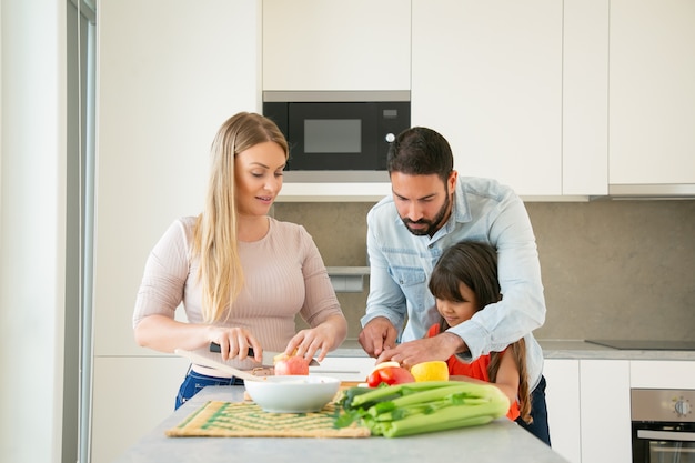 Mamá y papá enseñando a cocinar al niño. Pareja joven y su chica cortando frutas y verduras frescas para ensalada en la mesa de la cocina. Concepto de estilo de vida o nutrición saludable