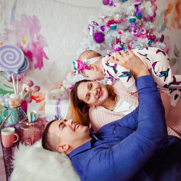 Mamá y papá se inclinan mutuamente a tender mentira con su pequeña hija bajo el árbol de Navidad rosa