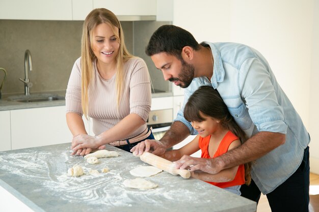 Mamá y papá positivos enseñando a su hija a enrollar la masa en la mesa de la cocina con harina desordenada. Pareja joven y su chica horneando bollos o pasteles juntos. Concepto de cocina familiar