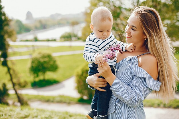 mamá de pelo largo en un vestido azul de pie en un parque solar y sosteniendo a su bebé