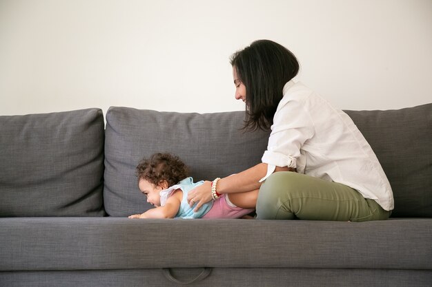 Foto gratuita mamá de pelo negro feliz abrazando a su hija linda en el sofá gris. vista lateral. concepto de paternidad e infancia