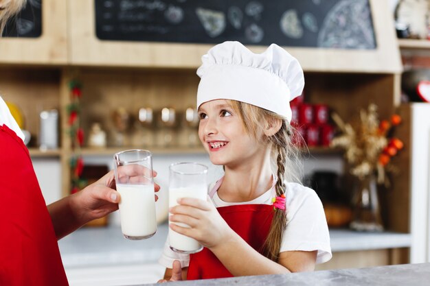 Mamá y pequeña hija encantadora se divierten bebiendo leche en la mesa en una cocina acogedora