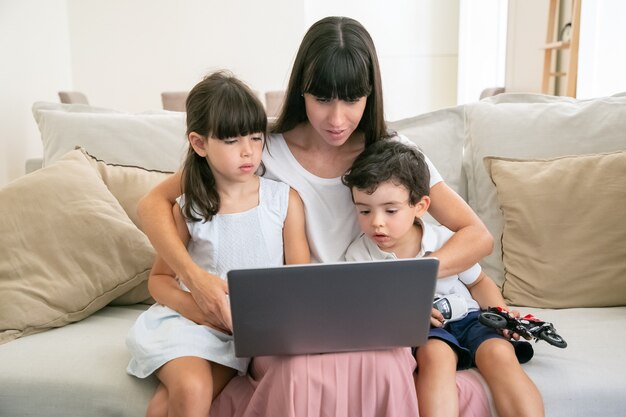 Mamá seria abrazando a niños preocupados mientras ven una película triste o de terror en la computadora portátil en casa