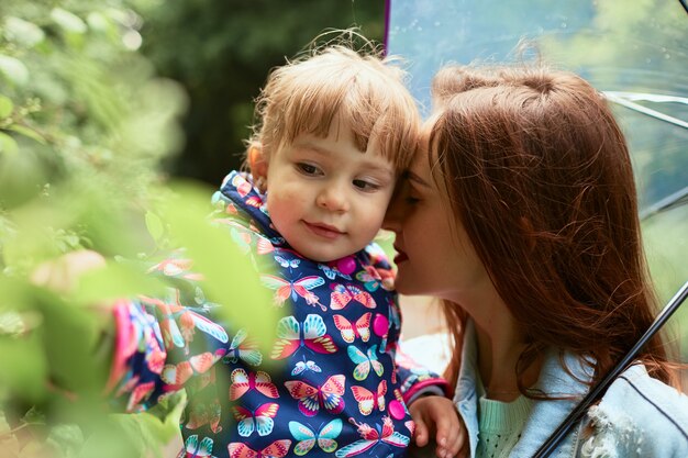 Mamá sostiene a su pequeña hija en manos de pie bajo el paraguas en el parque