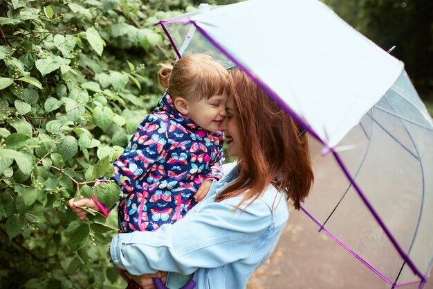 Mamá sostiene a su pequeña hija en manos de pie bajo el paraguas en el parque