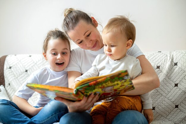 Mamá y sus dos hijos están leyendo un libro en casa en un sofá. Feliz y sonriente