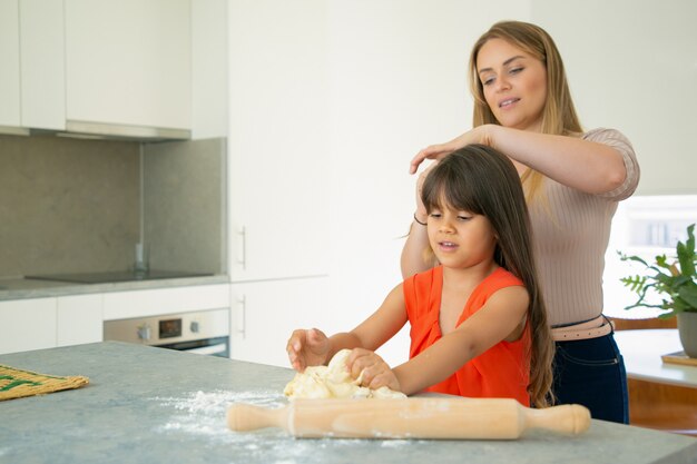 Mamá trenza el cabello largo de sus hijas mientras la niña hace masa en la cocina. Madre e hijo horneando juntos. Concepto de cocina familiar