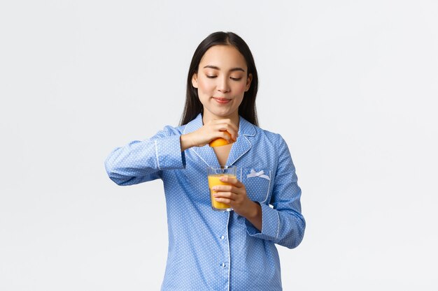 Mañana, estilo de vida activo y saludable y concepto de hogar. Chica asiática bastante saludable en pijama preparar jugo de naranja, exprimir naranja en vaso y sonriendo complacido, de pie fondo blanco