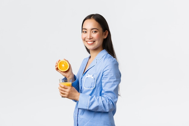 Mañana, estilo de vida activo y saludable y concepto de hogar. Perfil o hermosa chica asiática sana en pijama azul exprimiendo jugo de naranja en vaso y sonriendo feliz, comenzando el día a la derecha.
