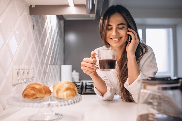 Mañana de la mujer con teléfono, croissant y café en la cocina.