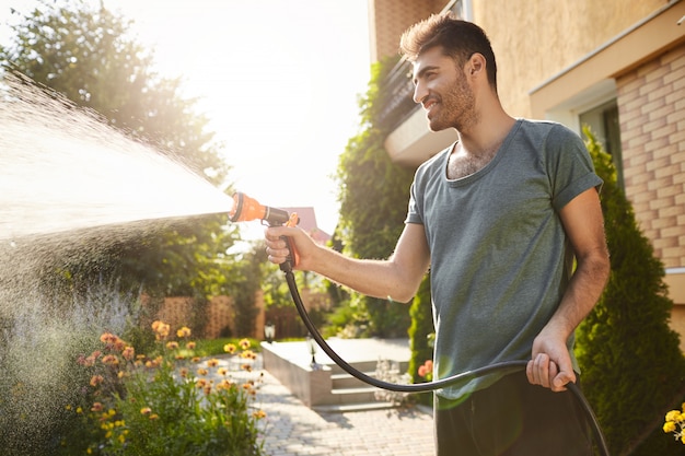 Foto gratuita mañana de verano en casa de campo. retrato de joven atractivo hombre barbudo de piel bronceada en camiseta azul sonriendo, regando las plantas con manguera, trabajando en el jardín.