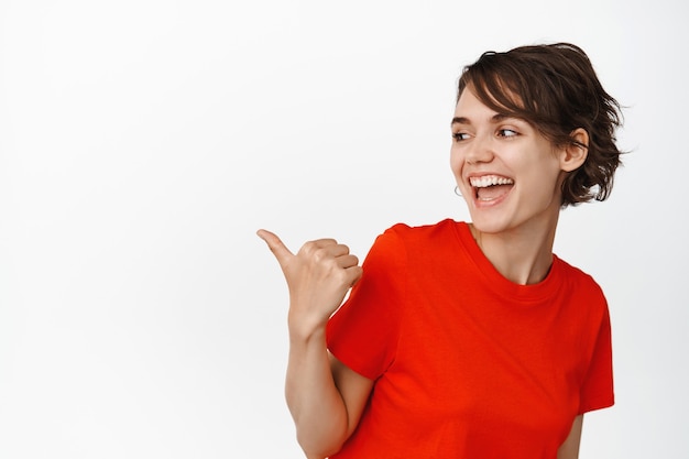 De esta manera. Hermosa chica moderna riendo, apuntando con el pulgar hacia la izquierda y mirando el espacio de la copia con una sonrisa feliz en blanco