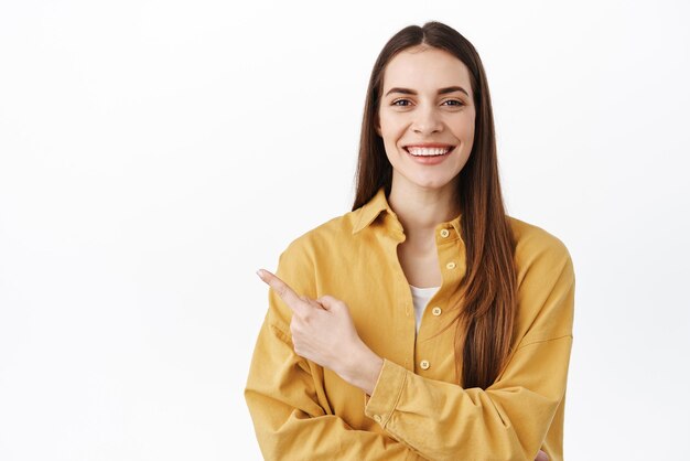 De esa manera, una modelo femenina sonriente y atractiva muestra la dirección señalando con el dedo a un lado y luciendo complacida recomendando una pancarta que muestra el logotipo de pie sobre un fondo blanco