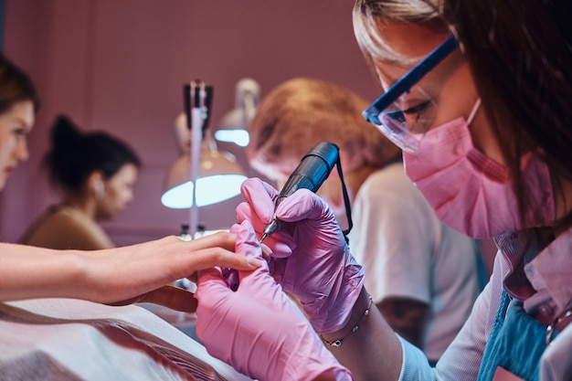 Una manicurista talentosa y enfocada está trabajando en su propio lugar de trabajo en un salón de belleza ocupado. Ella está usando una máscara protectora.