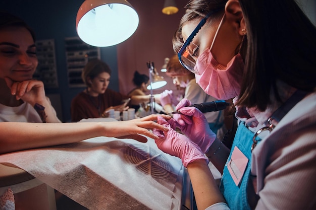 Una manicurista talentosa y enfocada está trabajando en su propio lugar de trabajo en un salón de belleza ocupado. Ella está usando una máscara protectora.