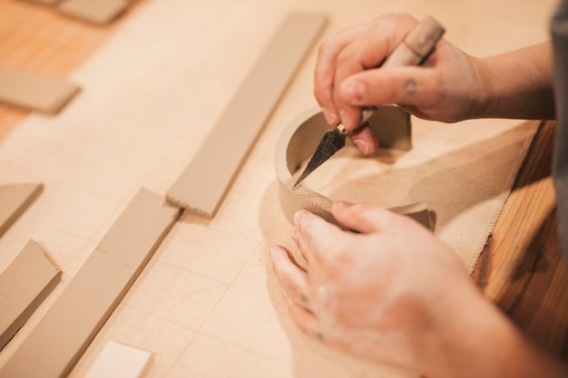 Mano de alfarero femenina grabando la arcilla con herramientas en mesa de madera