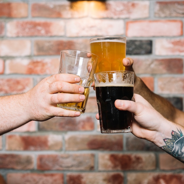 Foto gratuita la mano de un amigo tostado vasos de bebidas alcohólicas contra la pared de ladrillo