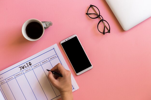 Mano con bolígrafo cerca de calendario, teléfono inteligente, taza de bebida y anteojos