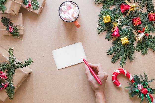 Mano con bolígrafo cerca de papel cerca de cajas presentes, ramita de navidad y taza