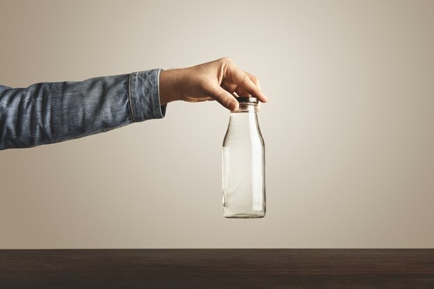 Mano brutal en chaqueta de jeans sostiene una botella transparente de vidrio con agua potable limpia para tapa metálica negra sobre la mesa de madera roja, aislada en blanco