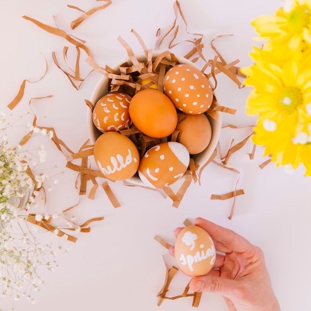 Mano cerca de conjunto de huevos de Pascua naranja en un tazón entre flores