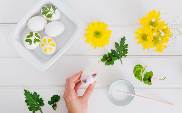 Mano cerca de los huevos de Pascua en el recipiente, flores, hojas y taza con líquido tinte