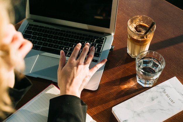 Mano de la empresaria que trabaja en la computadora portátil con un vaso de batido de chocolate y agua en el escritorio de madera