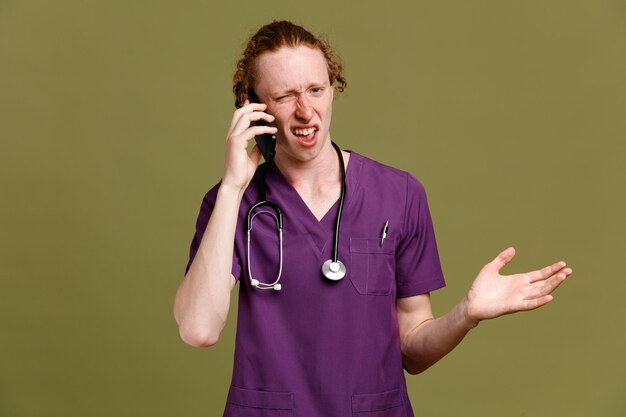 Mano extendida confundida habla por teléfono joven médico masculino vistiendo uniforme con estetoscopio aislado sobre fondo verde