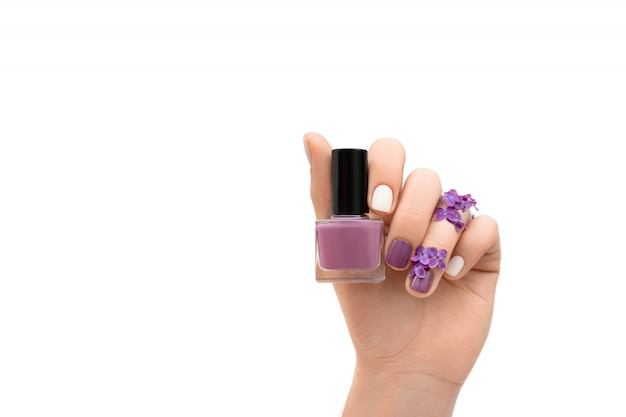 Mano femenina adornada con las flores de la lila que sostienen la botella púrpura del esmalte de uñas aislada en el fondo blanco. Concepto de primavera.