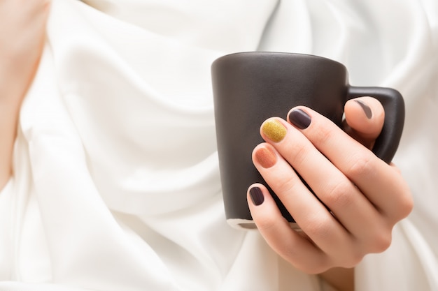 Mano femenina con diseño de uñas marrón con taza negra.