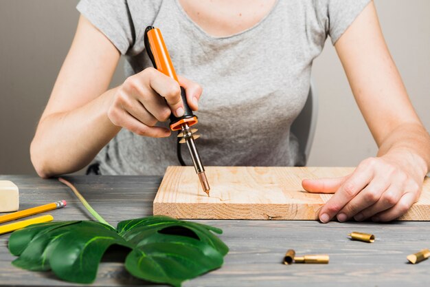 Foto gratuita mano femenina que corta la forma de madera dura usando la máquina que suelda cerca de la hoja de la monstera en la tabla