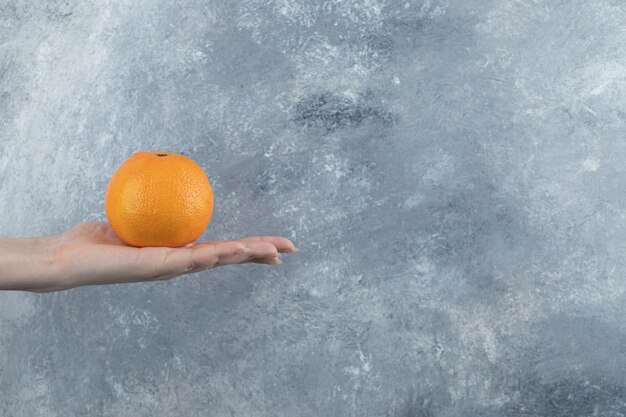 Mano femenina que sostiene una sola naranja en la mesa de mármol.