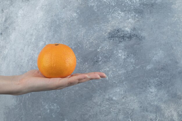 Mano femenina que sostiene una sola naranja en la mesa de mármol.