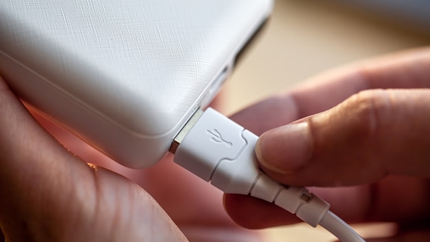 Una mano femenina sostiene un cable USB blanco y un banco de energía blanco, cargando el teléfono con un banco de energía, de cerca.