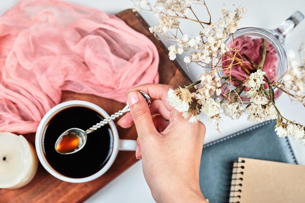 Mano holdong una taza de café sobre tabla de madera con velas, tela ponk y flores.