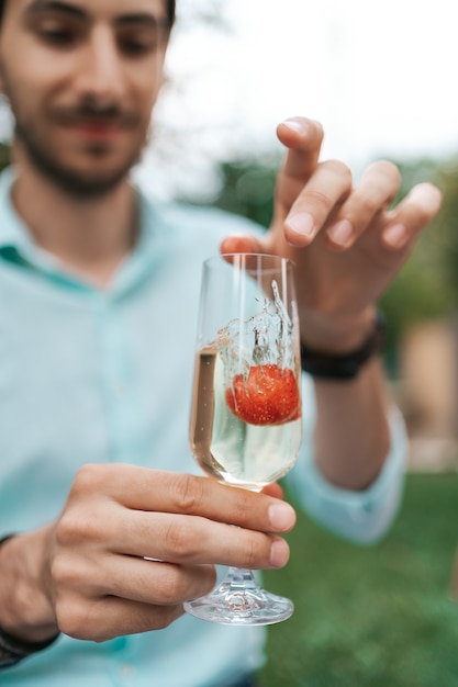 La mano del hombre deja caer una fresa en un vaso con vino espumoso. Hermosa vida, celebración