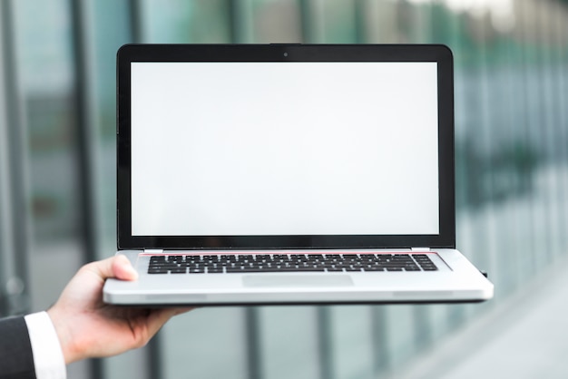 La mano de un hombre de negocios que muestra la computadora portátil con la pantalla blanca en blanco contra el contexto borroso