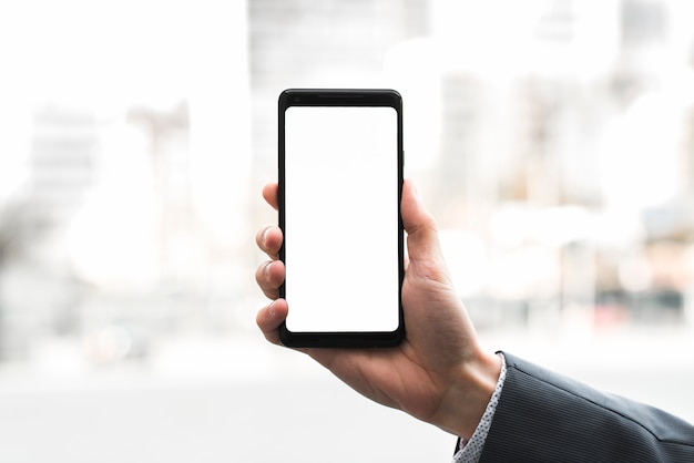 La mano de un hombre de negocios que muestra el teléfono móvil contra fondo borroso