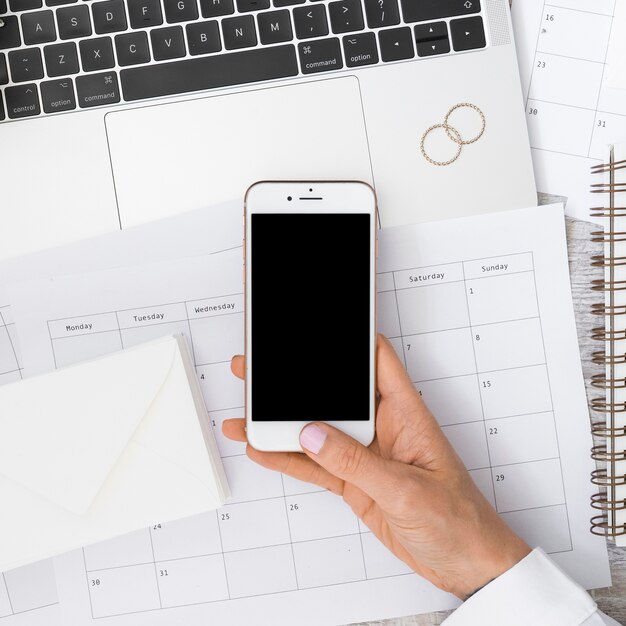 La mano del hombre de negocios que sostiene smartphone sobre el calendario con el sobre; portátil y anillos de boda