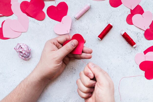 La mano del hombre que hace la guirnalda de papel roja y rosada de la forma del corazón con el hilo en el fondo blanco