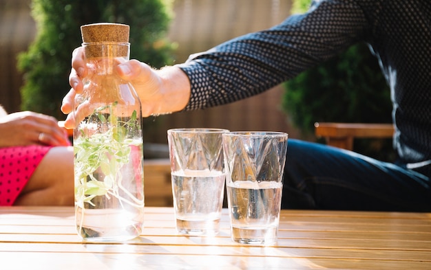 La mano del hombre que sostiene la botella herbaria de cristal de la bebida con dos vidrios en la tabla de madera