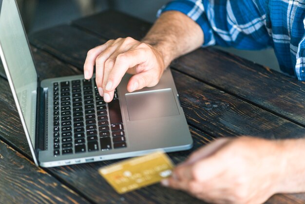 La mano del hombre que sostiene la tarjeta de crédito que mecanografía en la computadora portátil sobre el escritorio de madera