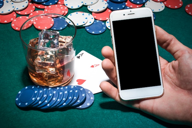 Mano del hombre que sostiene el teléfono móvil sobre la mesa de póquer con vaso de whisky
