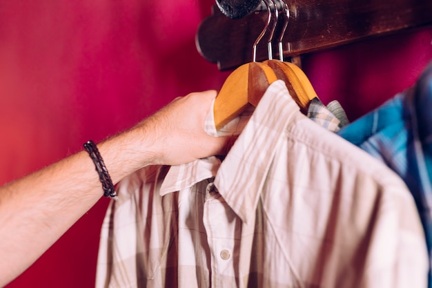 La mano del hombre que toma la camisa de la percha del gancho del estante en la pared roja