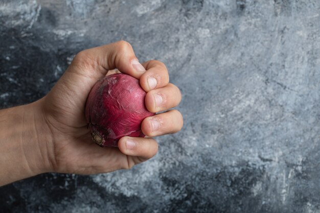 Mano de hombre sosteniendo una cebolla morada sobre un fondo gris.