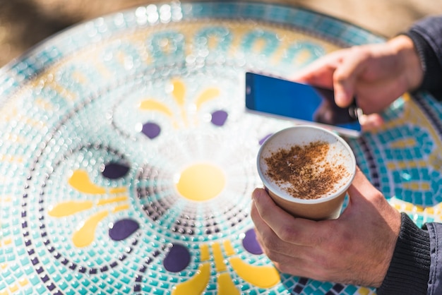 La mano del hombre sosteniendo tazas de capuchino espolvoreado con chocolate en polvo usando el teléfono móvil