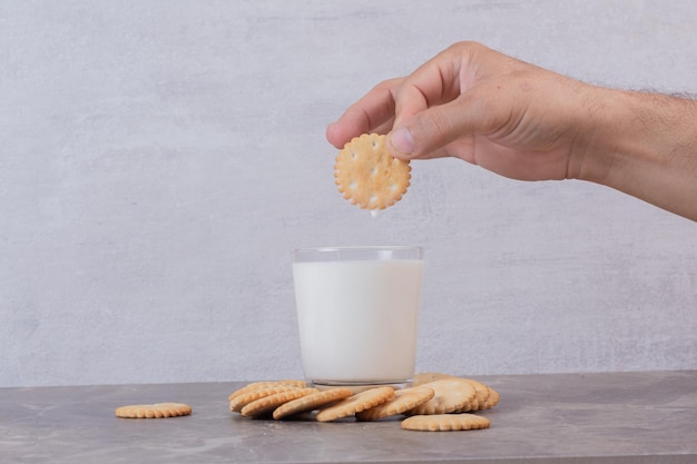 La mano del hombre sostiene una galleta encima de la leche en la mesa de mármol.